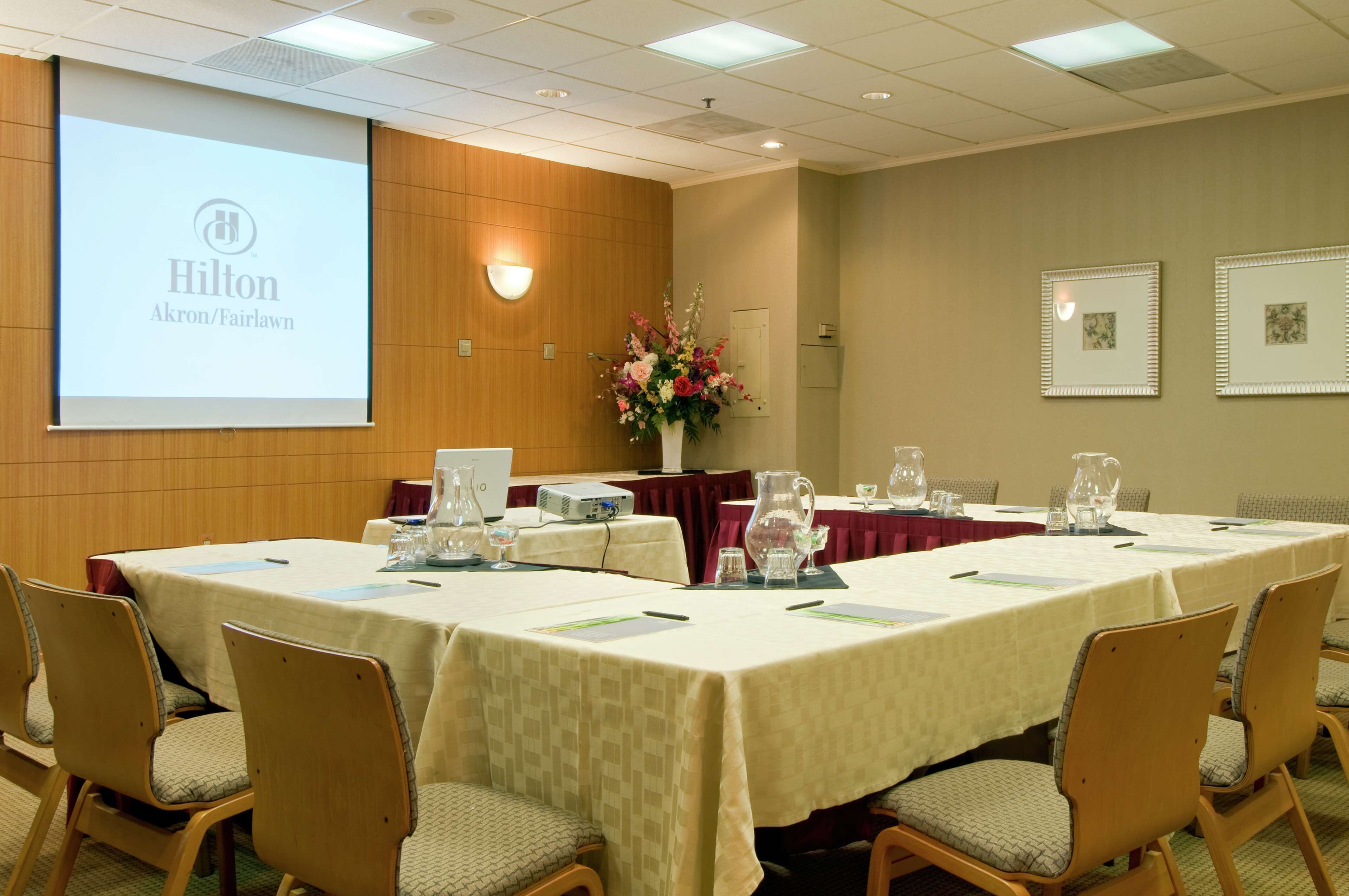 A conference room with a large screen displaying 'Hilton Akron/Fairlawn', a floral arrangement, and a table set for a meeting.