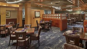 A hotel dining area with multiple tables and chairs, wooden paneling, and a patterned carpet.