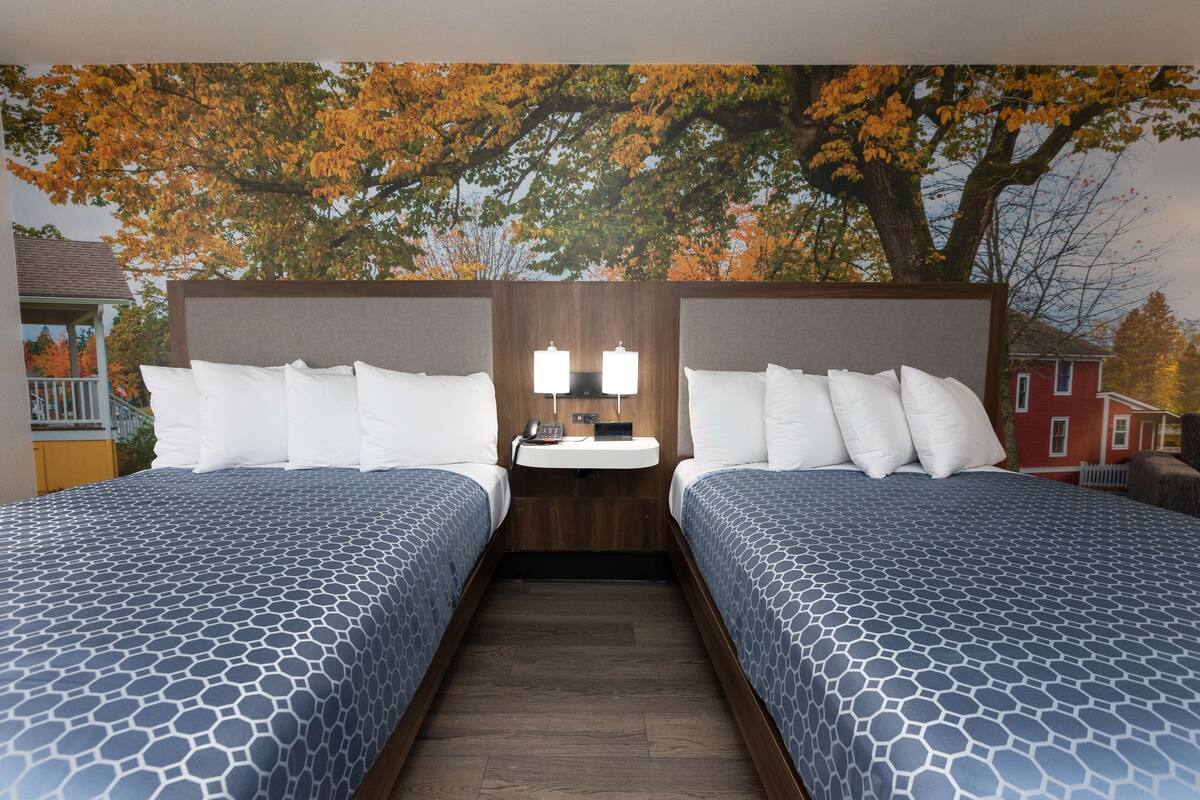A hotel room with two beds, a nightstand with a lamp, and a wall mural of autumn trees.