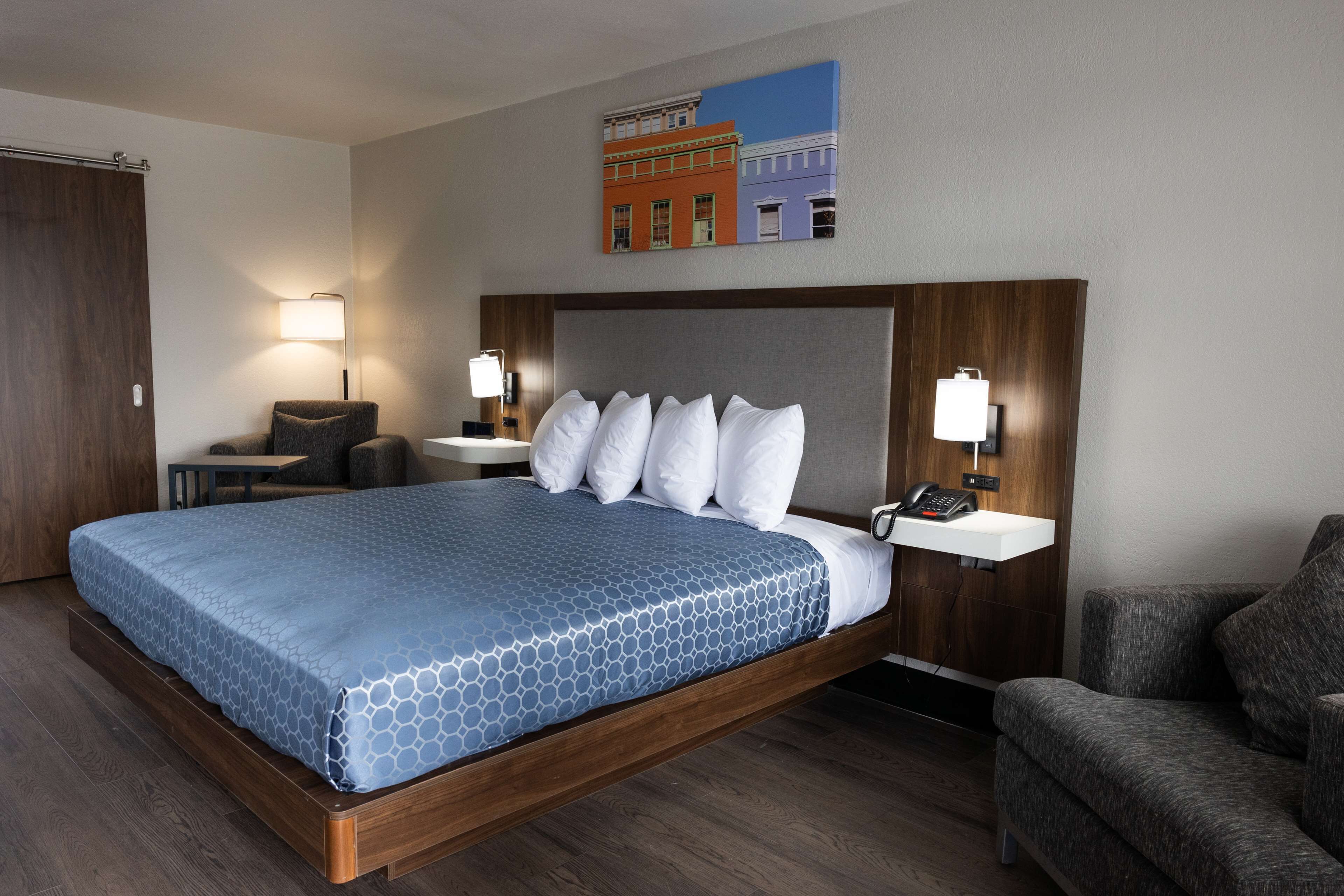 Un cuarto de hotel con una cama grande, dos mesitas de noche con lámparas, una silla y un cuadro en la pared.