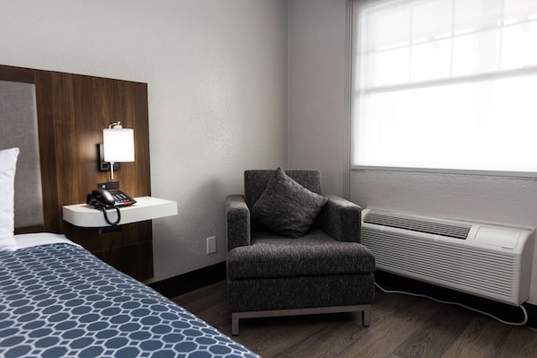 Habitación de hotel con cama, una silla, escritorio con teléfono, lámpara y ventana con vistas.