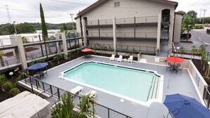 Una piscina en la azotea con varias sillas de descanso y sombrillas, rodeada por un edificio con techo rojo y múltiples balcones.