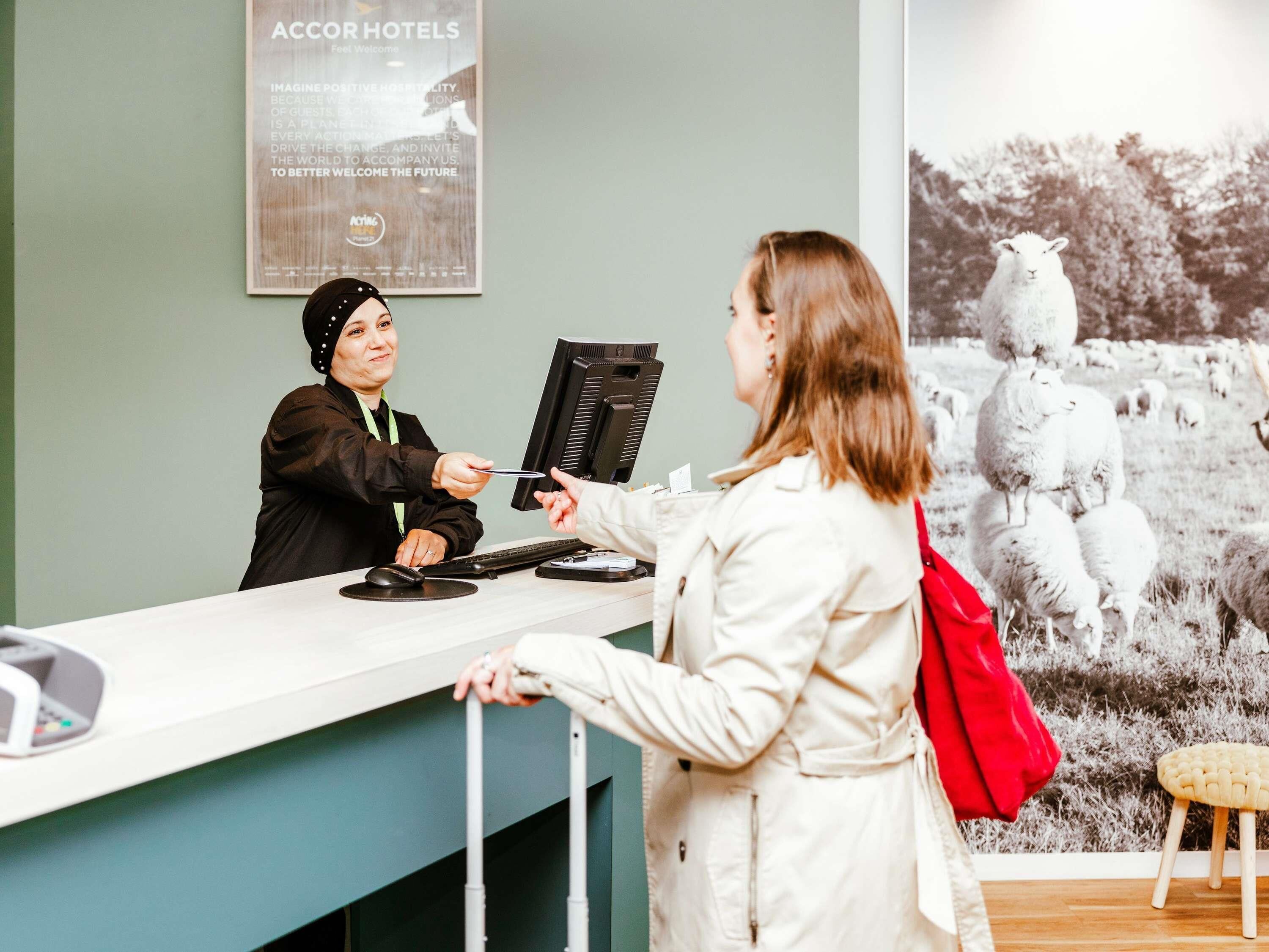 A hotel reception desk with a computer monitor, a keyboard, and a mouse. There is a poster on the wall and a photograph of sheep in the background.