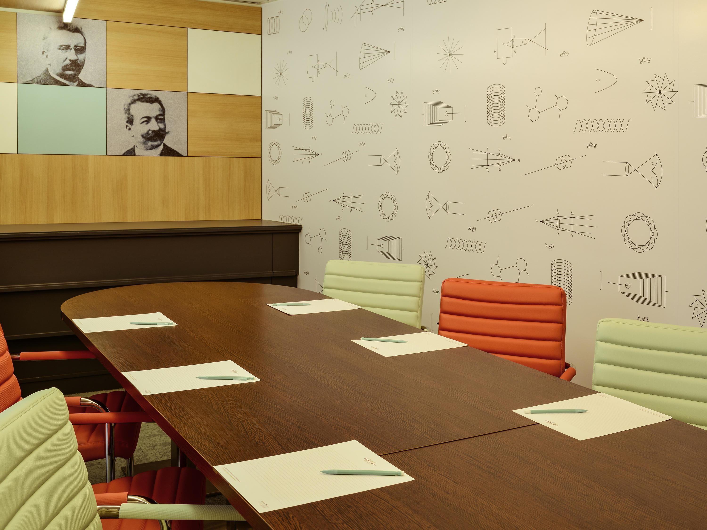 Una sala de conferencias con una mesa de madera, sillas coloridas y una pared con diagramas científicos.