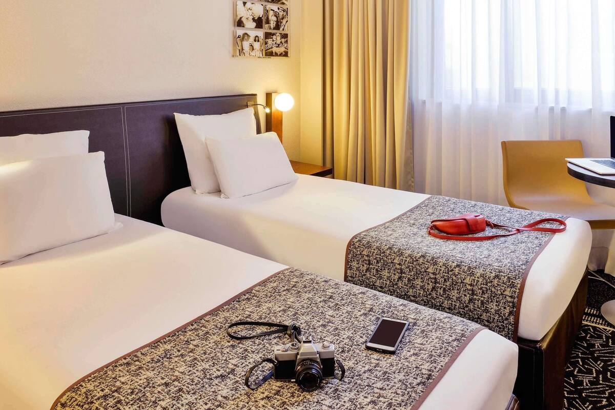 A hotel room with two beds, a desk, a chair, and a lamp.