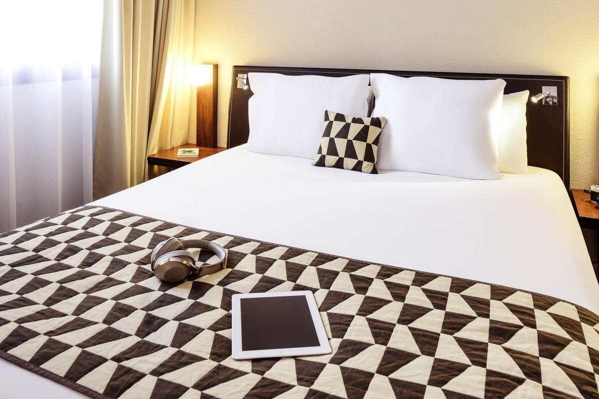 A hotel room with a neatly made bed, a bedside table with a lamp, and a window with curtains.