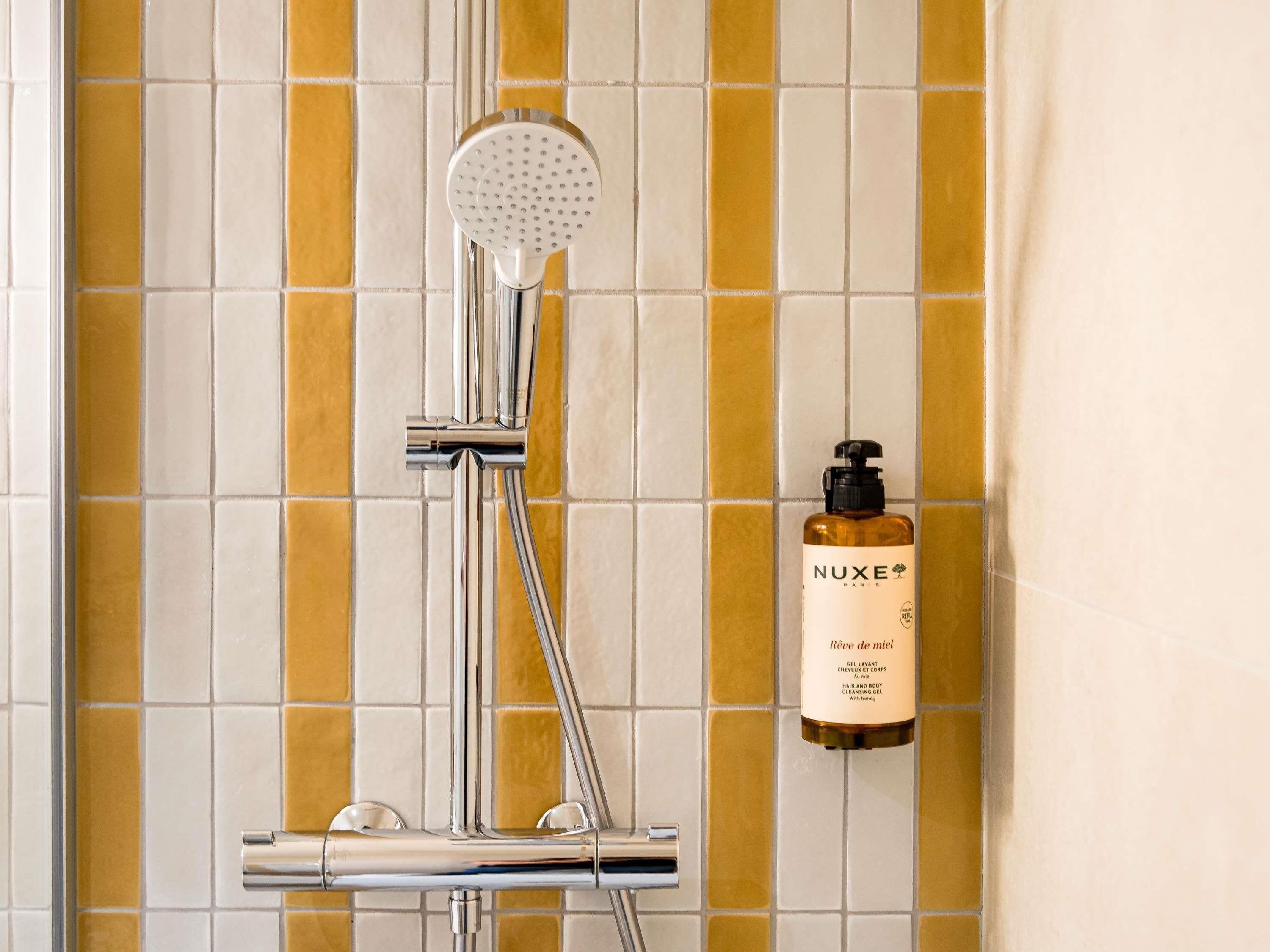 Una ducha con cabezal de ducha manual, una botella de gel limpiador para cabello y cuerpo Nuxe Paris fijada a la pared, y una pared de azulejos con un diseño amarillo y blanco.