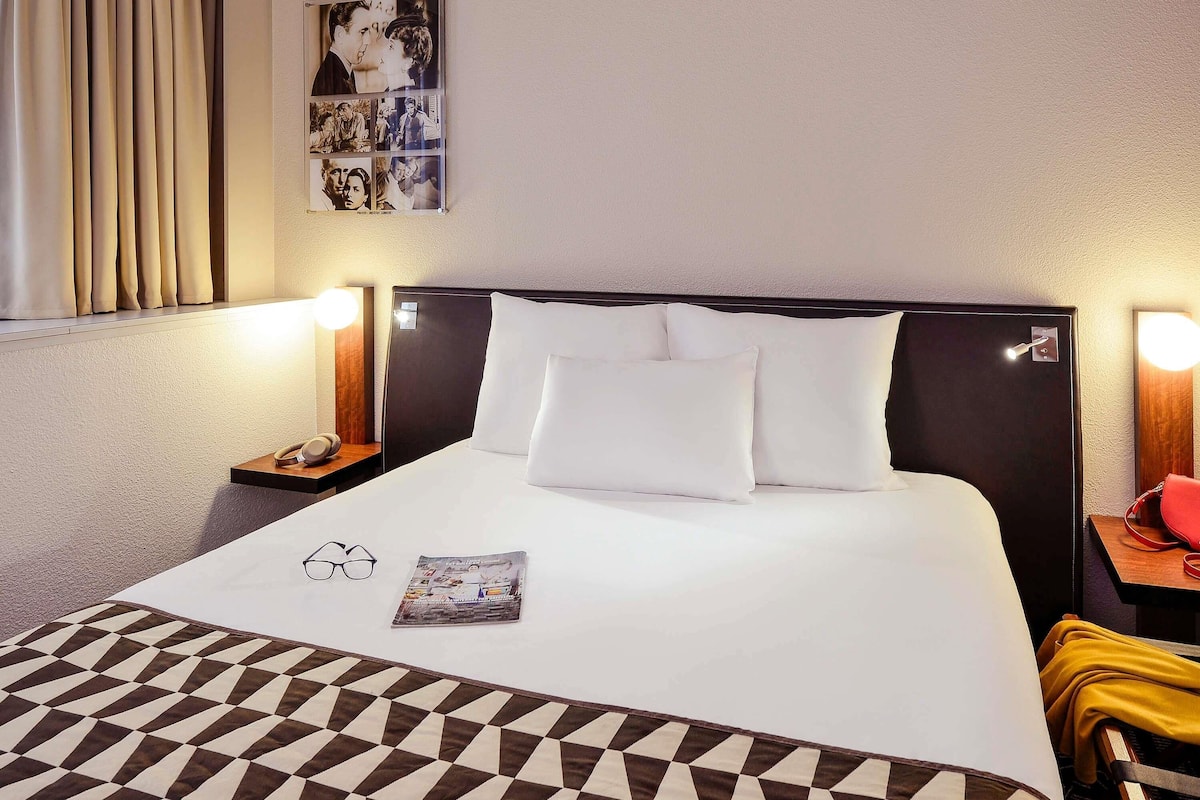 Une chambre d’hôtel avec un lit, des lampes de chevet, une table de chevet, une horloge murale, un magazine et un tapis à motifs.