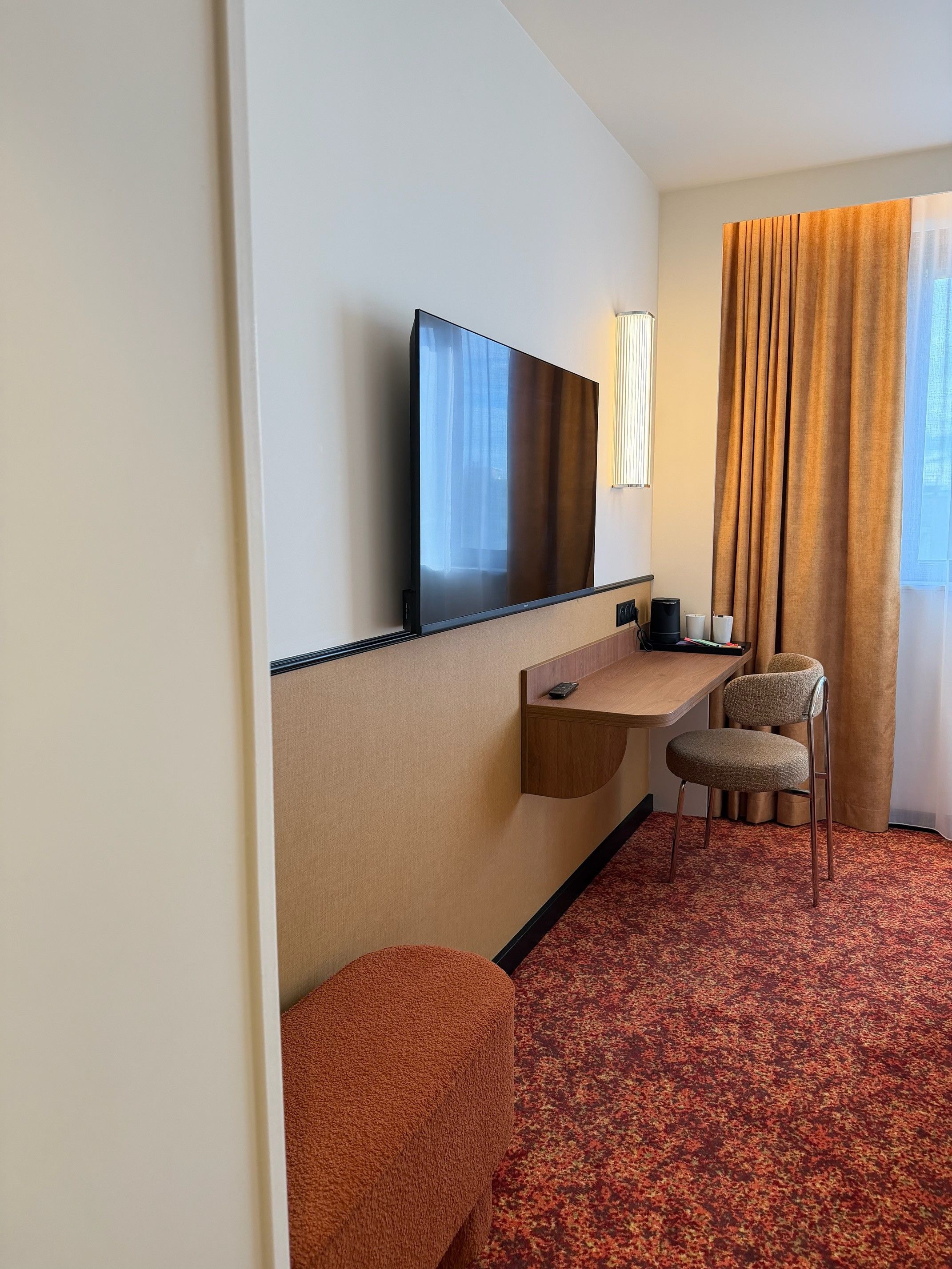 Et hotelværelse med et fladskærms TV på væggen, et lille træskrivebord med stol og et stort vindue med gardiner.