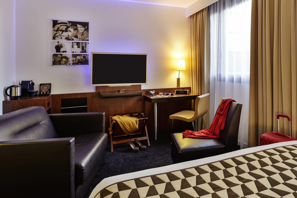 Habitación de hotel con cama, escritorio, silla, televisor y estante de pared con fotografías.