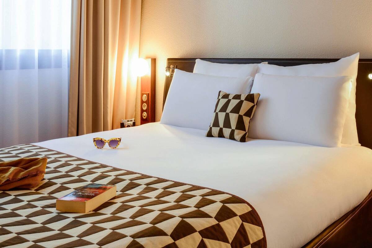 Une chambre d’hôtel avec un lit, un plaid à motifs, un livre et une paire de lunettes de soleil posées sur le lit.