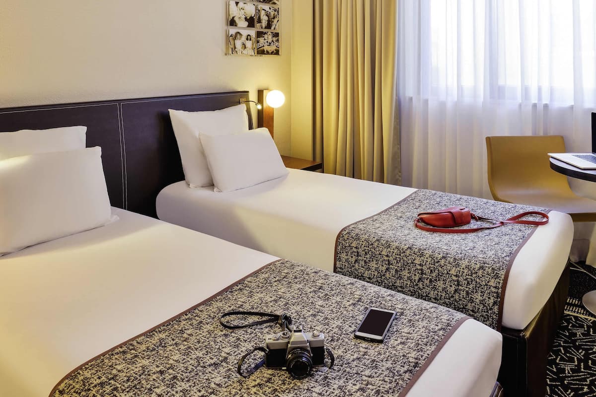 Quarto de hotel com duas camas, uma escrivaninha, uma cadeira, uma luminária e uma parede decorada com quadros.
