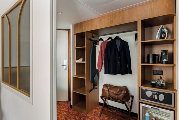 Habitación de hotel con vestidor, perchero con ropa colgada, maleta, cafetera y refrigerador.