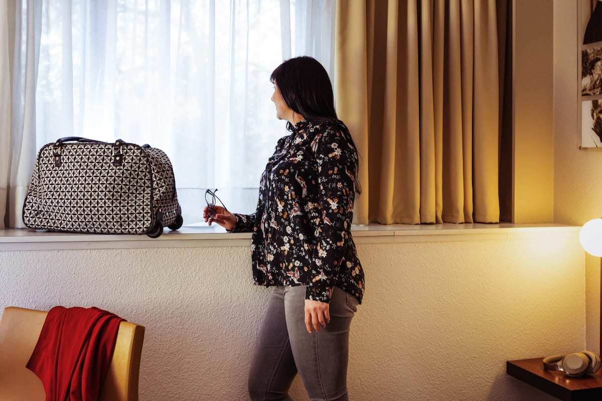 A room with a window, a suitcase, a chair with a red blanket, and a lamp.