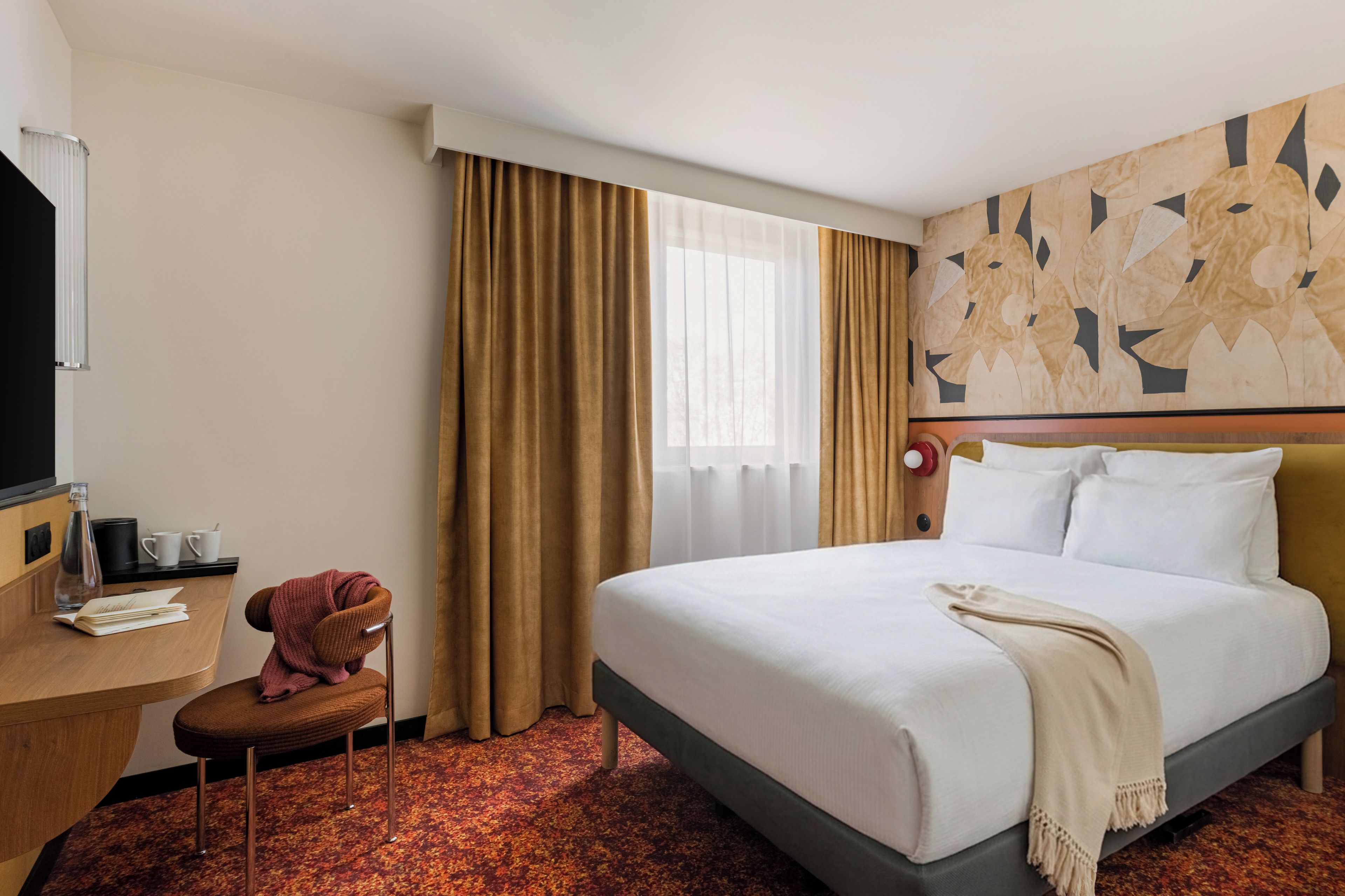Habitación de hotel con cama, escritorio, silla, ventana con cortinas y pared con diseño geométrico.