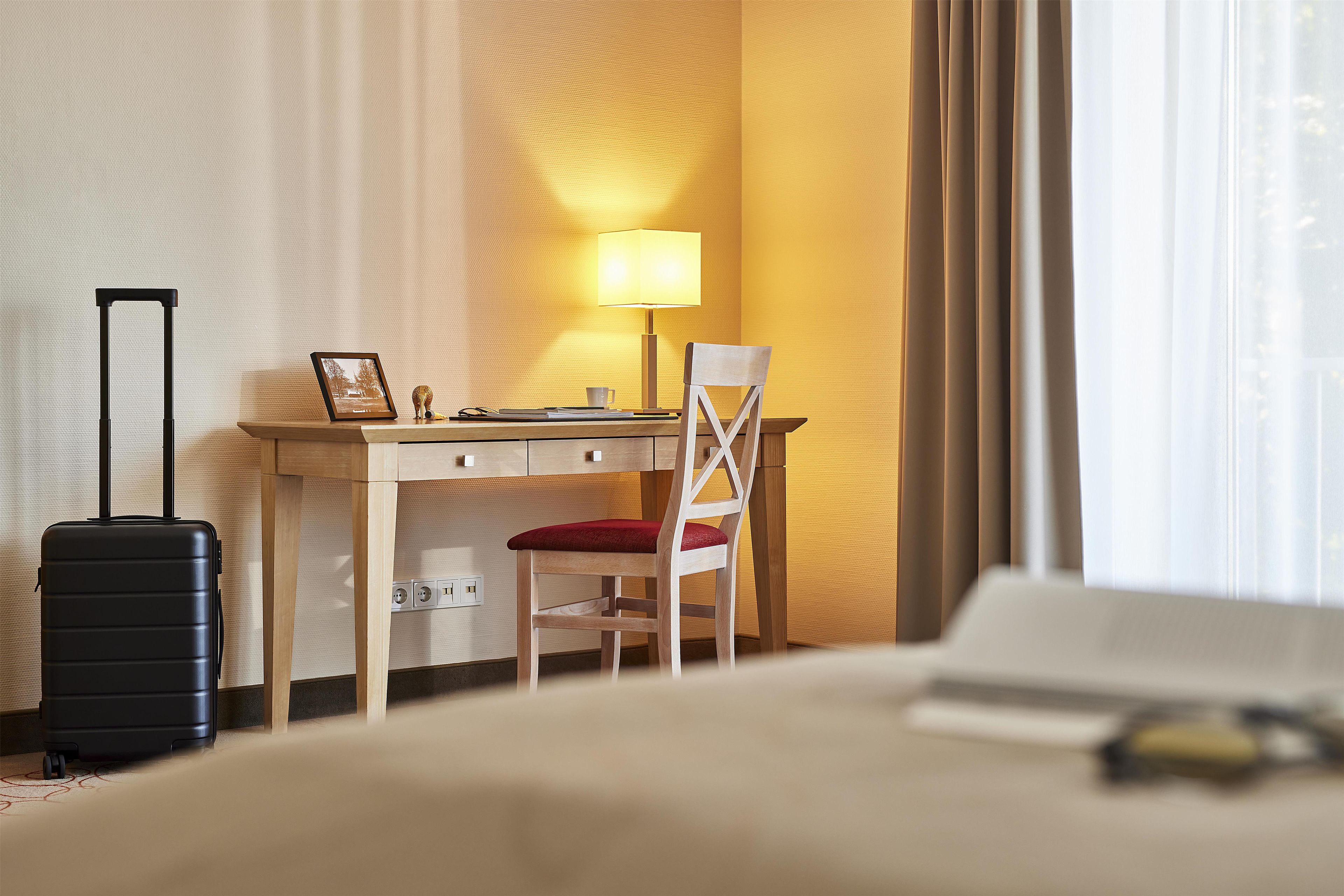 A hotel room with a bed, a desk with a chair, a lamp, and a suitcase.