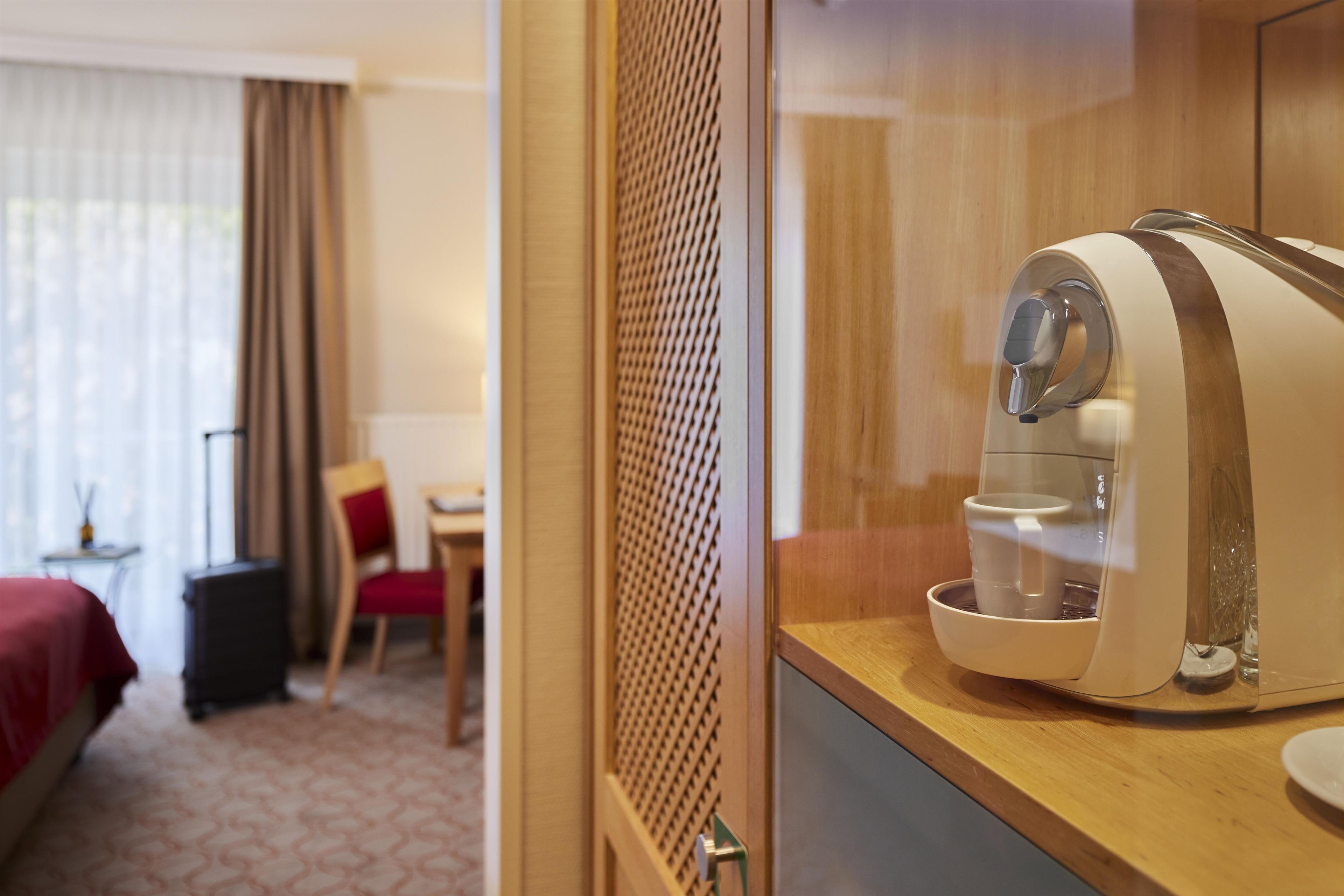 A hotel room with a coffee machine on a wooden counter, a bed with a red cover, a chair, and a small table with a lamp.