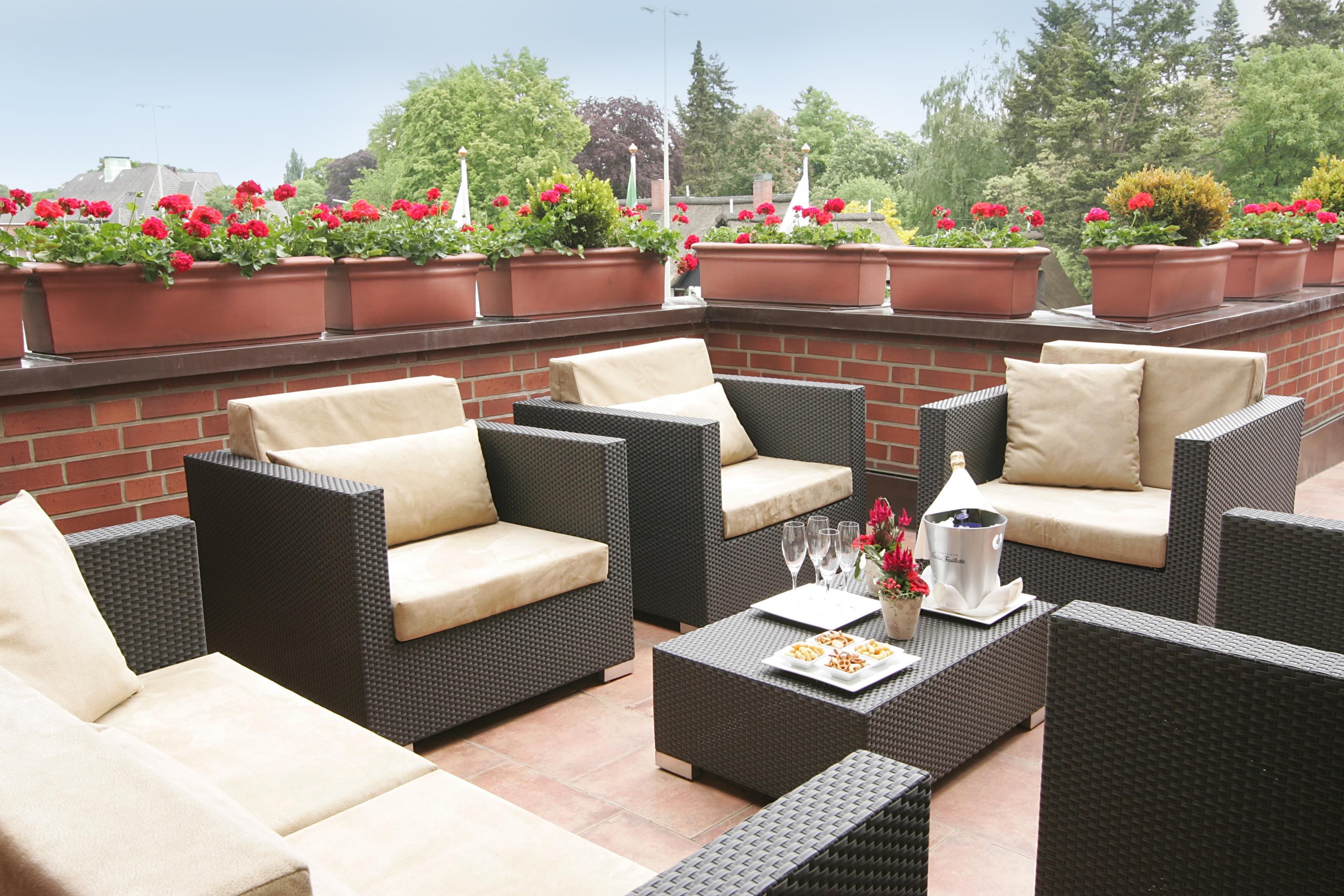 An outdoor patio with wicker furniture, a table with a bottle and glasses, and red flowers in planters.