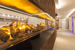Un pasillo con una vitrina de cristal que contiene rocas y una chimenea, una pared con una pintura de un paisaje urbano, y una puerta con una luz azul.