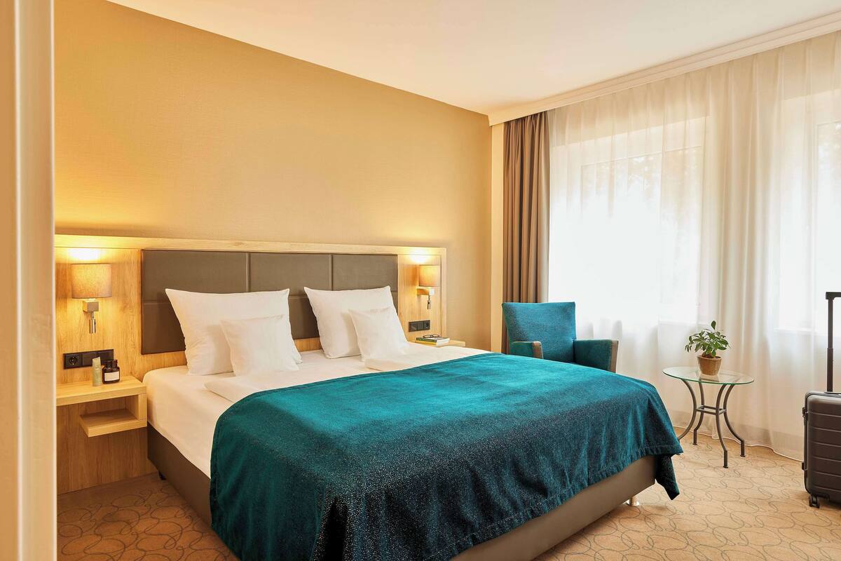 Une chambre d’hôtel avec un lit, des tables de chevet, une chaise, une petite table avec une plante et une valise.