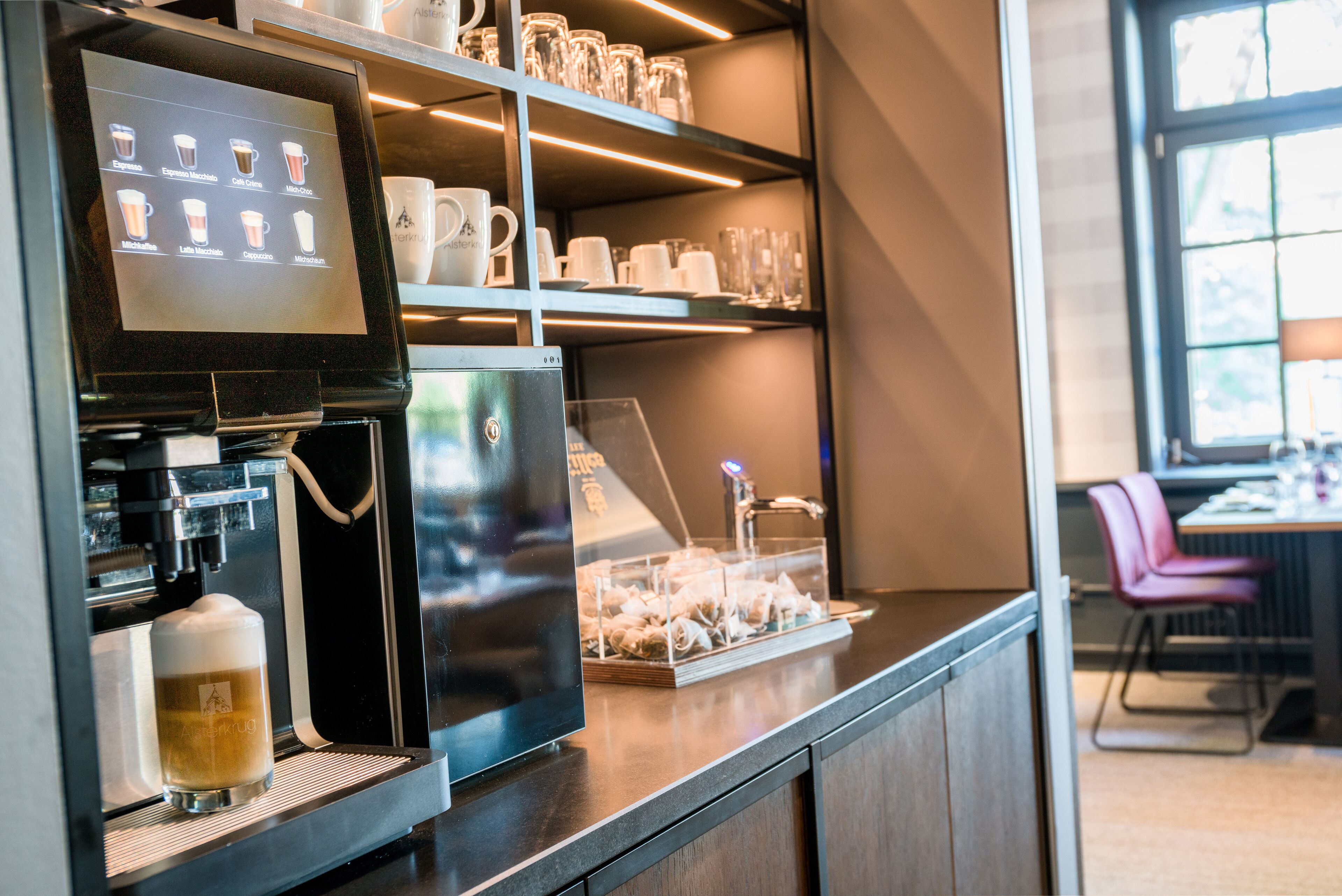 A coffee machine with a touchscreen menu, a glass display case with pastries, and shelves with cups and mugs.
