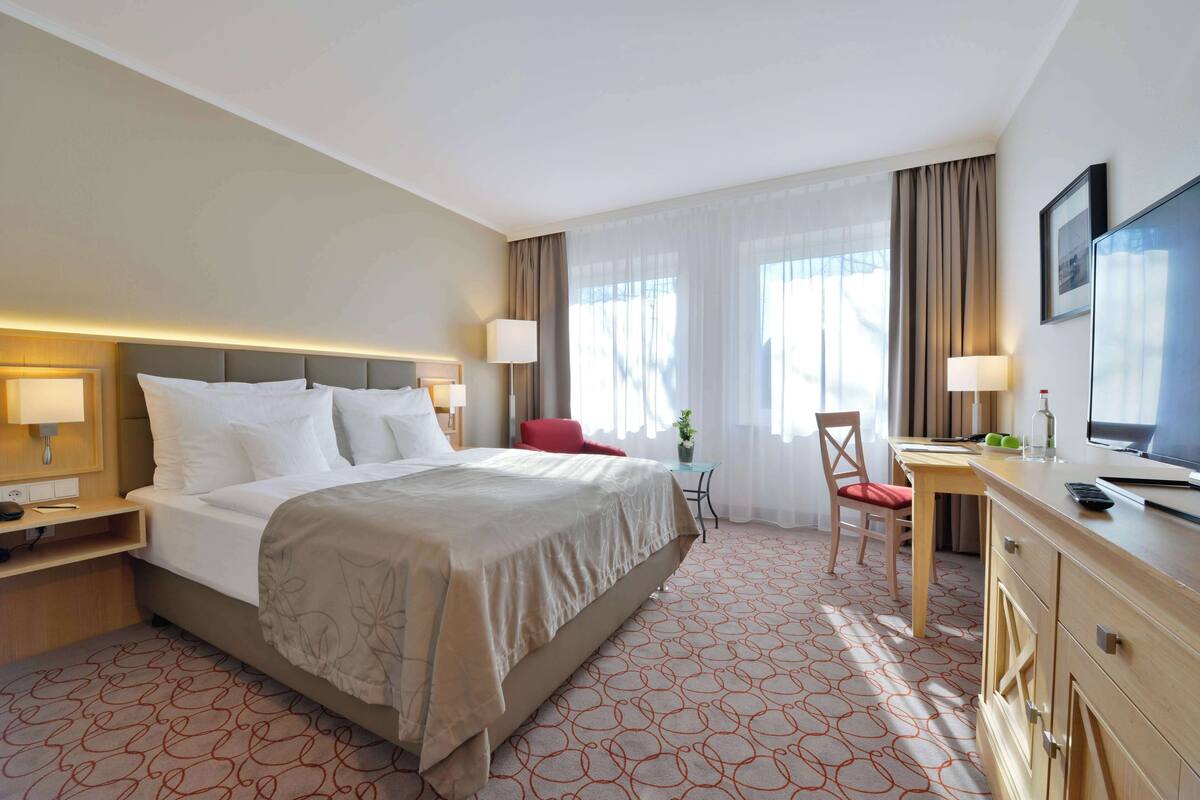 Une chambre d’hôtel avec un grand lit, un bureau avec une chaise, une télévision et une fenêtre avec des rideaux.