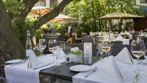 Un área de comedor al aire libre con mesas dispuestas para una comida, que incluye manteles blancos, copas de vino y cubiertos.