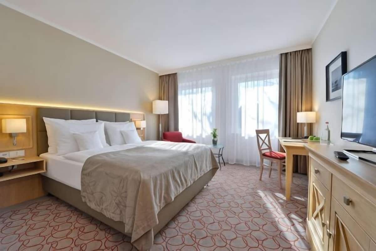 Una habitació d'hotel amb un llit, taules de nit, escriptori amb cadira, televisió i finestra amb cortines.