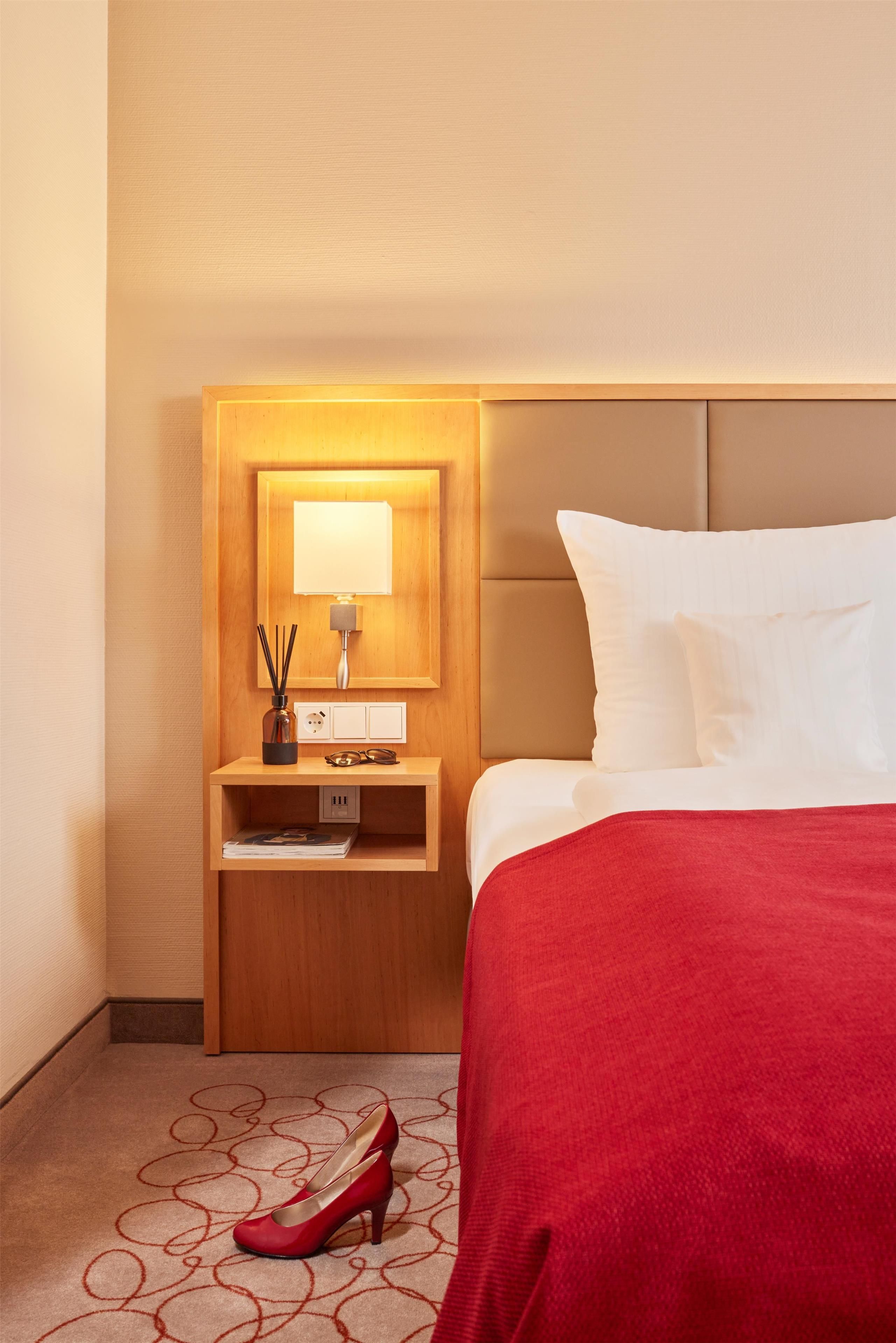 A hotel room with a neatly made bed, a bedside table with a lamp, and a pair of red shoes on the floor.