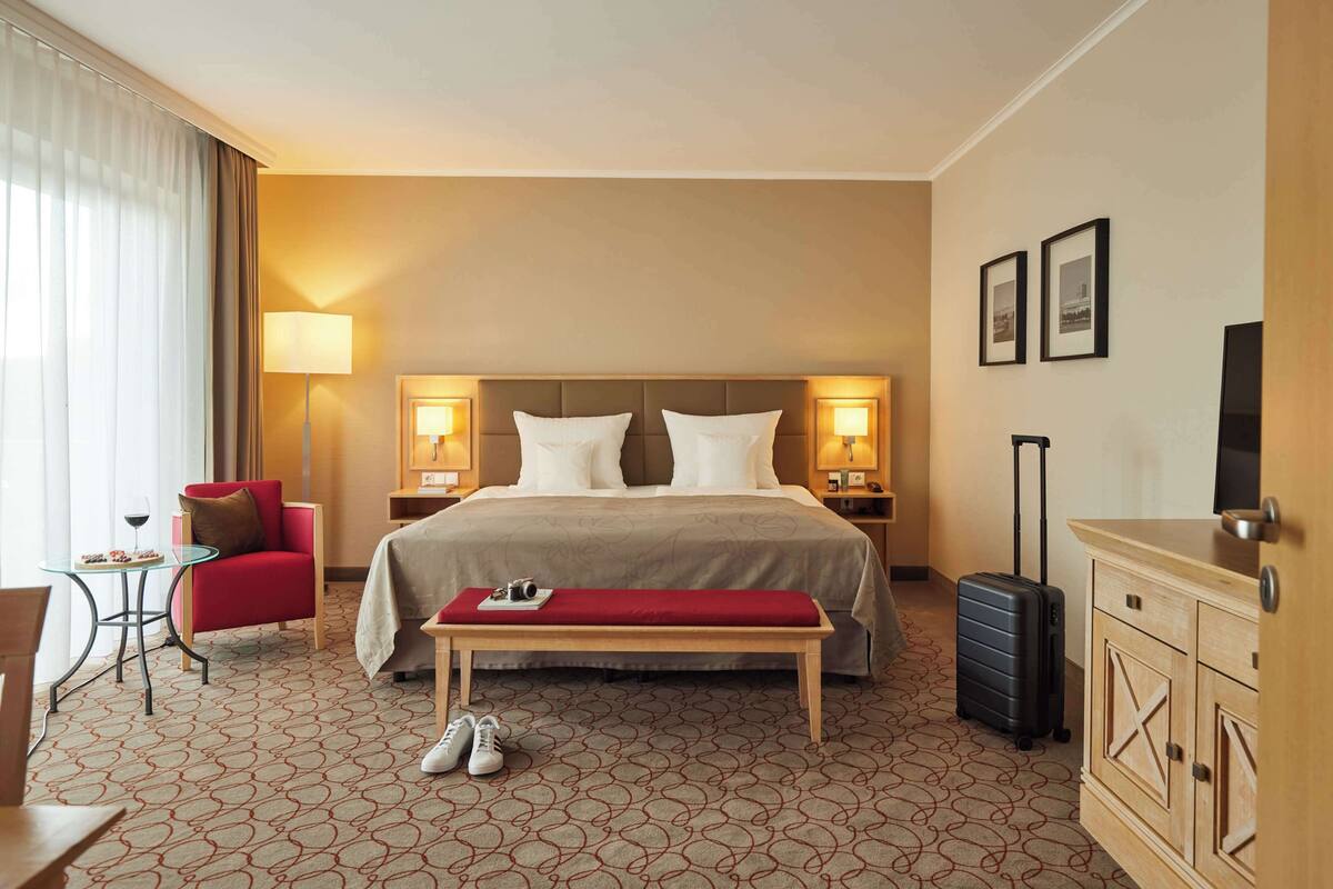 Une chambre d’hôtel avec un grand lit, deux lampes de chevet, un fauteuil rouge, une petite table avec un verre à vin, un banc, une valise et un téléviseur posé sur une commode en bois.