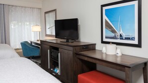 Habitación de hotel con cama, escritorio con televisor, silla, lámpara, cafetera y un cuadro en la pared.