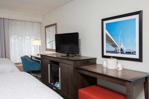 Habitación de hotel con cama, escritorio con televisor, silla, lámpara, cafetera y un cuadro en la pared.