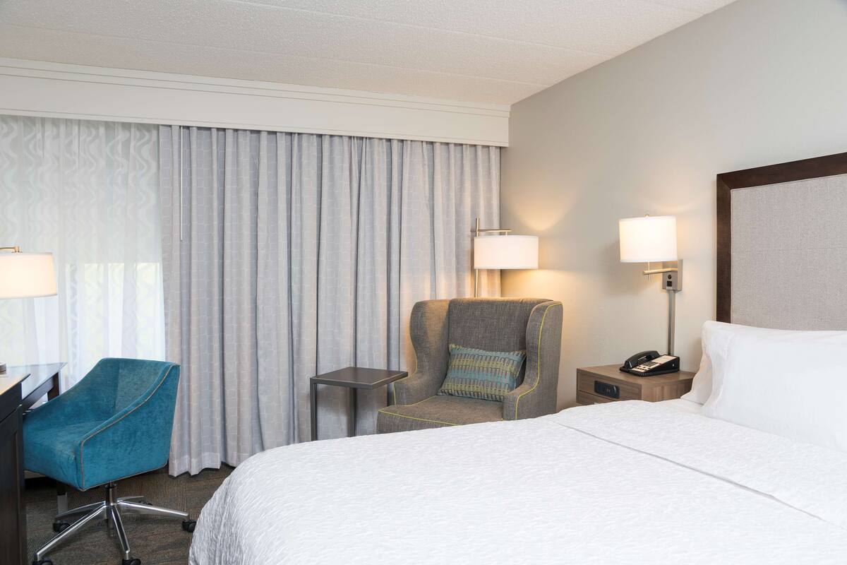 Una habitación de hotel con una cama, un escritorio con silla, una mesita y dos lámparas de noche.