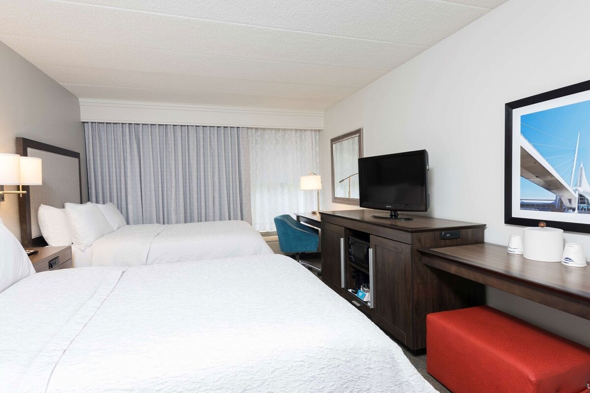 Une chambre d’hôtel avec deux lits, un bureau avec une télévision, un banc rouge et une grande fenêtre donnant sur un pont.