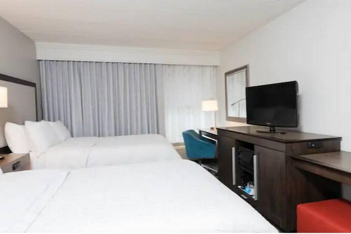 Habitación de hotel con dos camas, un escritorio, un televisor de pantalla plana, una silla y un amplio ventanal con cortinas.
