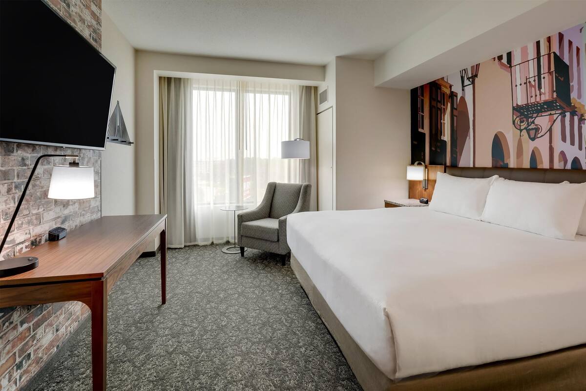 Une chambre d’hôtel avec un grand lit, un bureau avec une lampe, une chaise, un téléviseur fixé au mur en briques et une fenêtre avec des rideaux.