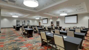 A conference room with multiple tables and chairs, a patterned carpet, ceiling lights, and a projector screen.