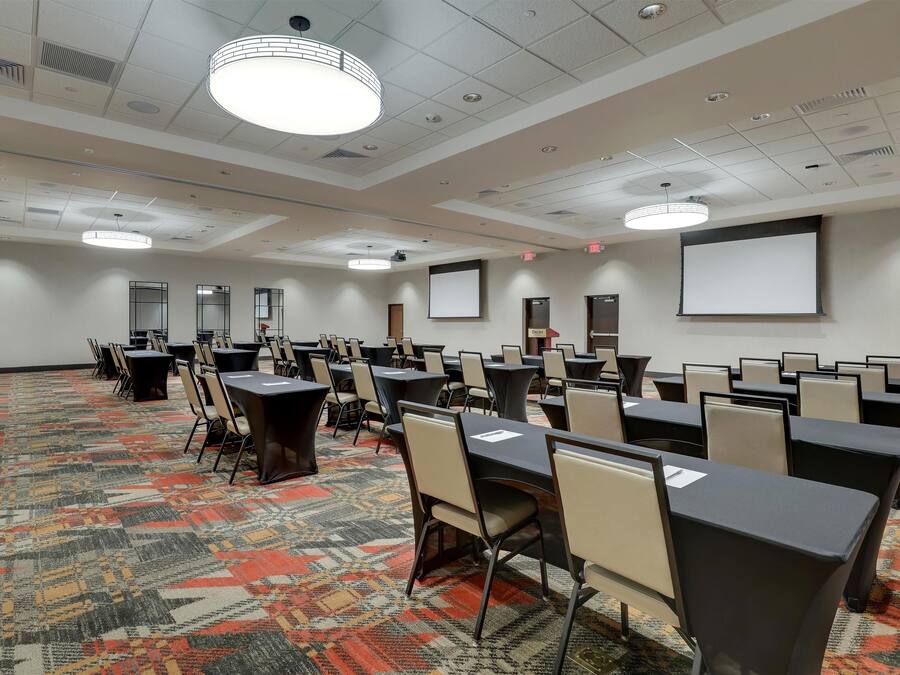 A conference room with multiple tables and chairs, a patterned carpet, ceiling lights, and a projector screen.