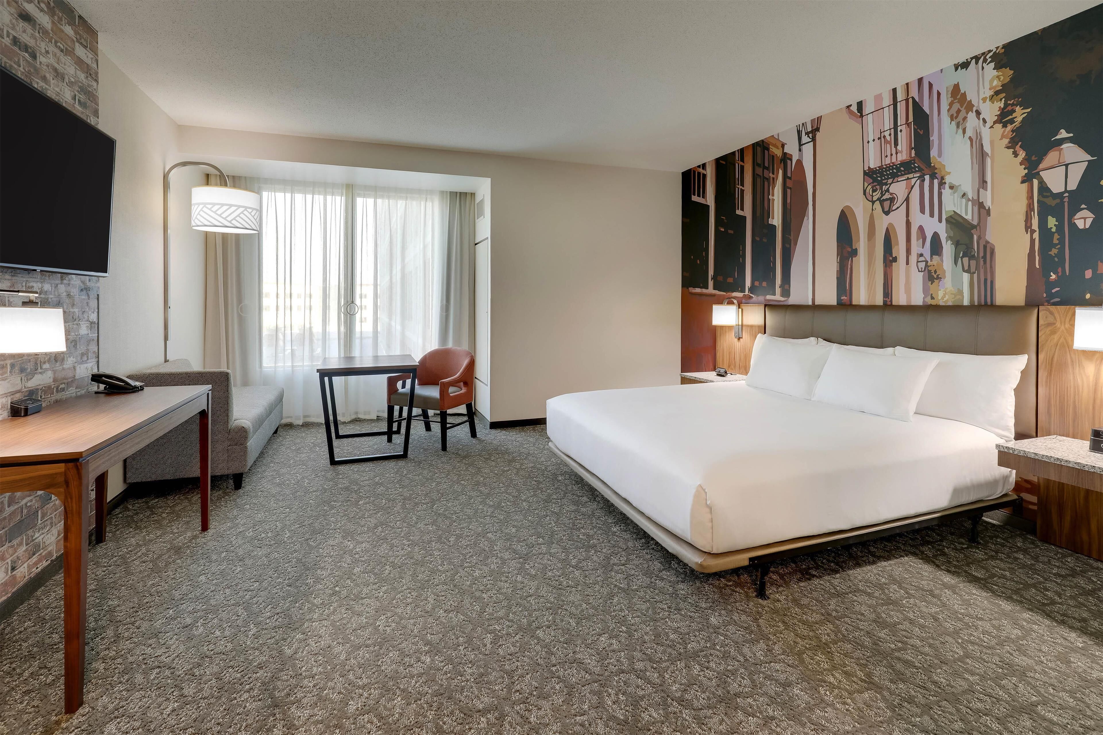 A hotel room with a large bed, a desk with a chair, a TV mounted on the wall, and a window with curtains.