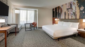 A hotel room with a large bed, a desk with a chair, a TV mounted on the wall, and a window with curtains.