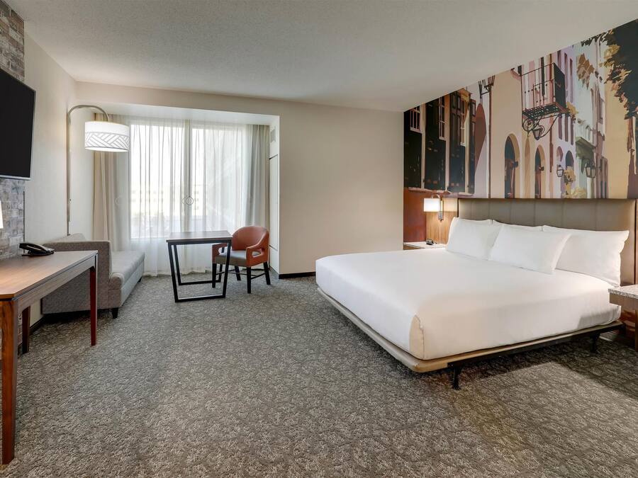 A hotel room with a large bed, a desk with a chair, a TV mounted on the wall, and a window with curtains.