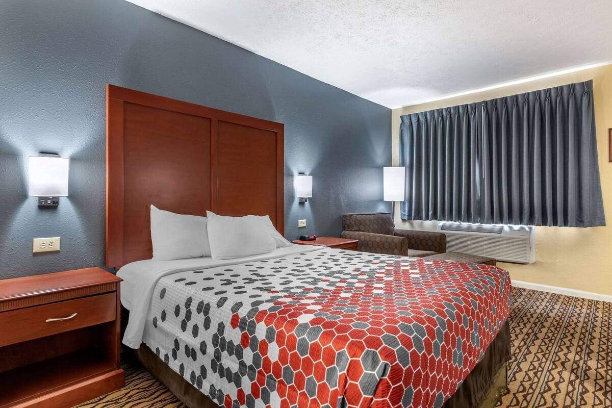 Quarto de hotel com cama, mesas de cabeceira, uma cadeira, escrivaninha, abajur e janela com cortinas.