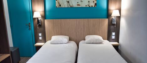 A hotel room with two single beds, a teal-colored door, a wooden headboard, and a wall-mounted painting of flowers.