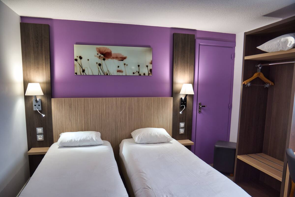 A hotel room with two single beds, a wooden headboard, purple walls, a painting, a wardrobe, and bedside lamps.