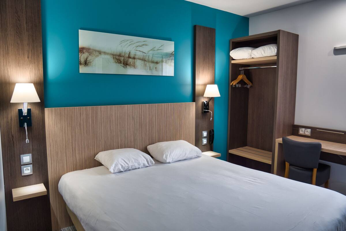 A hotel room with a large bed, two bedside lamps, a wooden headboard, a wall-mounted artwork, a wooden wardrobe, and a desk with a chair.
