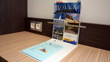 Un mostrador de recepción de hotel con un expositor de folletos que muestra un folleto de 'Rouen Normandie', una guía de la ciudad y una tarjeta de bienvenida.