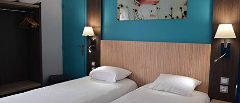 Habitación de hotel con dos camas individuales, cabecero de madera, pared de acento color turquesa, un cuadro y dos lámparas fijadas a la pared.