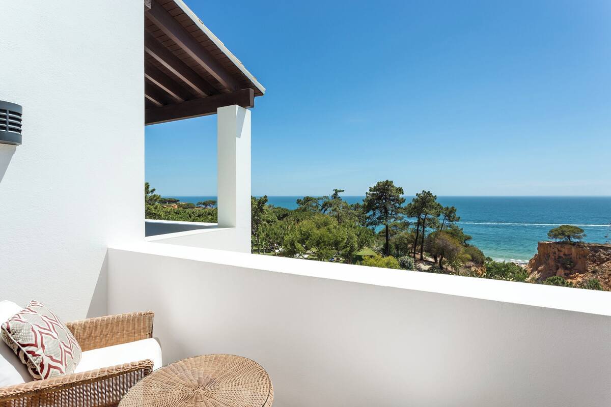 A balcony with wicker furniture, a white wall, and a view of the sea and trees.