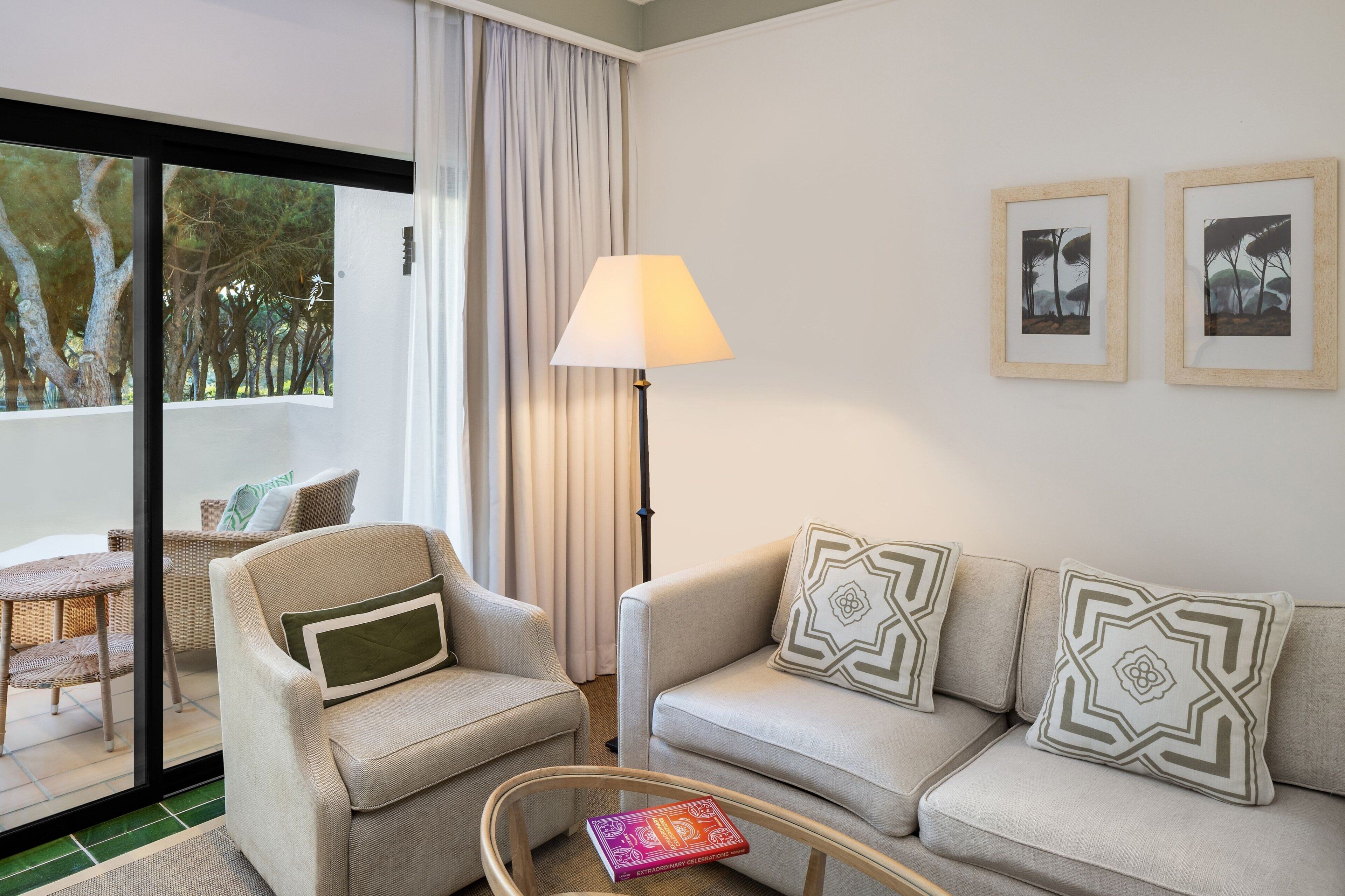 Ein modernes Wohnzimmer mit Sofa, Sessel und Stehlampe. Ein Glastür öffnet sich zu einem Außenbereich mit Blick auf Bäume.