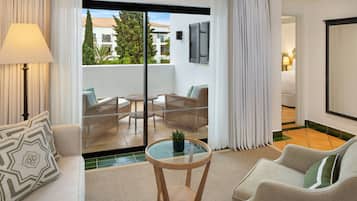 Habitación de hotel moderna con balcón, sofá, una mesa redonda con planta y vistas a edificios y zonas verdes exteriores.